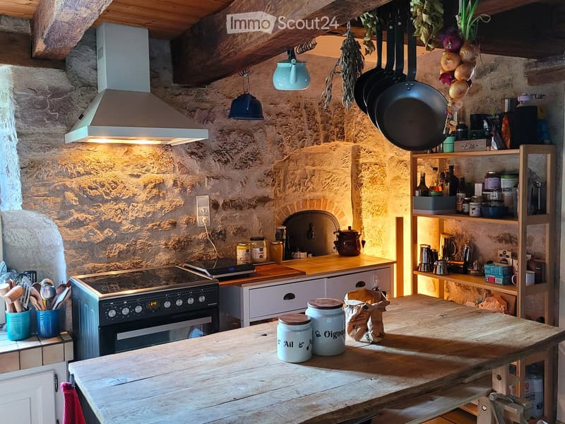 Rustic kitchen with stone walls, wooden beams, and a central wooden table. Features an oven, shelves with jars and bottles, hanging frying pans, and a rustic wooden countertop.