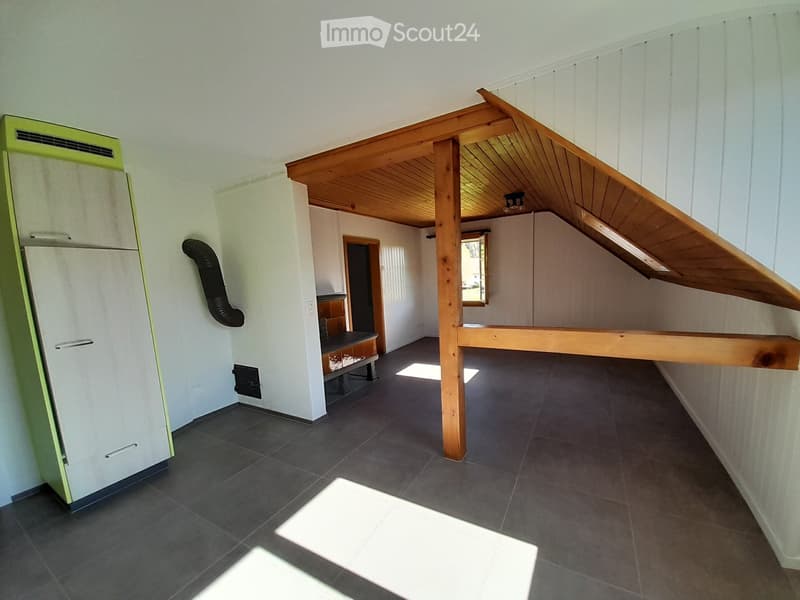 A modern, open-concept room with a slanted wooden ceiling and exposed wooden beams. Features include a refrigerator, a small table with chairs, and a built-in loft bed area.