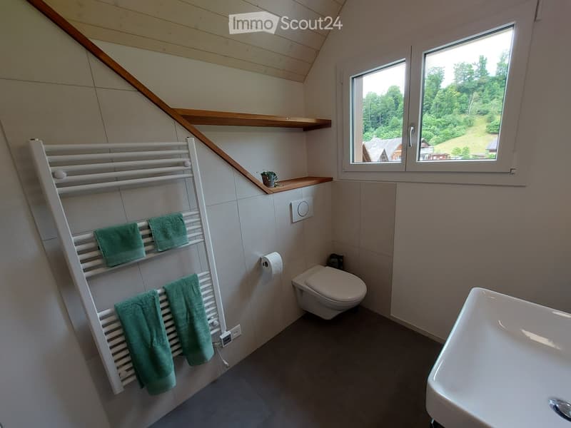 Modern bathroom with a toilet, sink, towel rack with green towels, and a small shelf with a plant. The room has large windows offering a view of greenery outside.