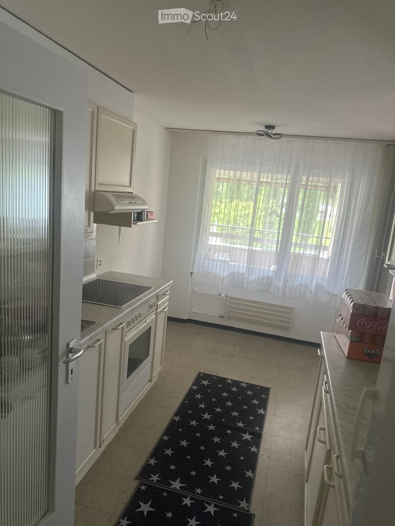 Modern kitchen with white cabinets, stainless steel appliances, and a black star-patterned rug. A sliding glass door leads to a balcony with a view of greenery.