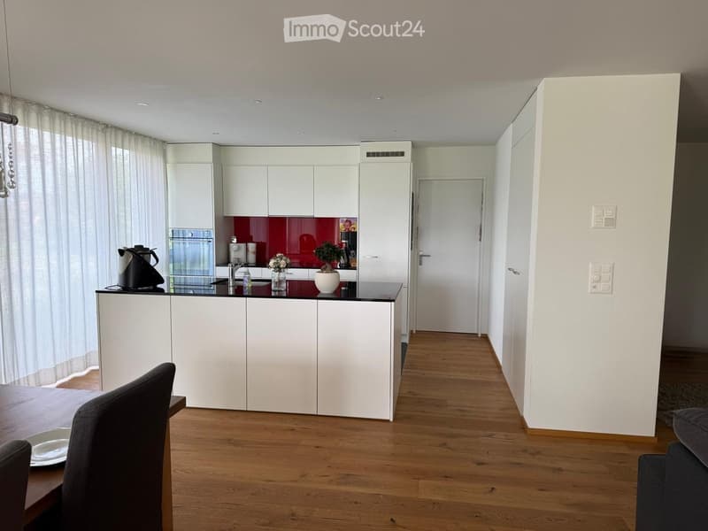 Modern kitchen with white cabinets, a black countertop, and a wooden dining area. Features include a teapot, vase with flowers, potted plant, and a curtained window.