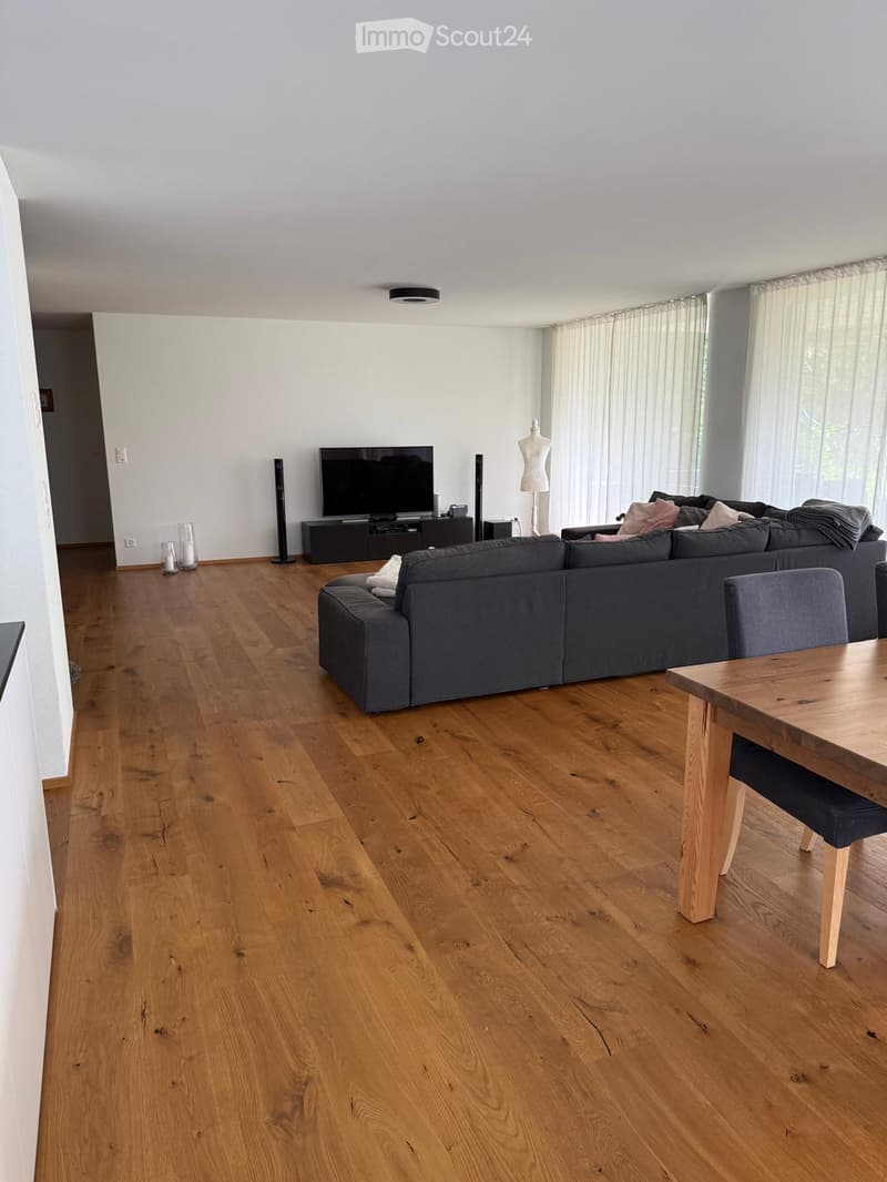 Modern living room with wooden flooring, large windows with curtains, a grey sofa, a wooden dining table, and a television on a stand.