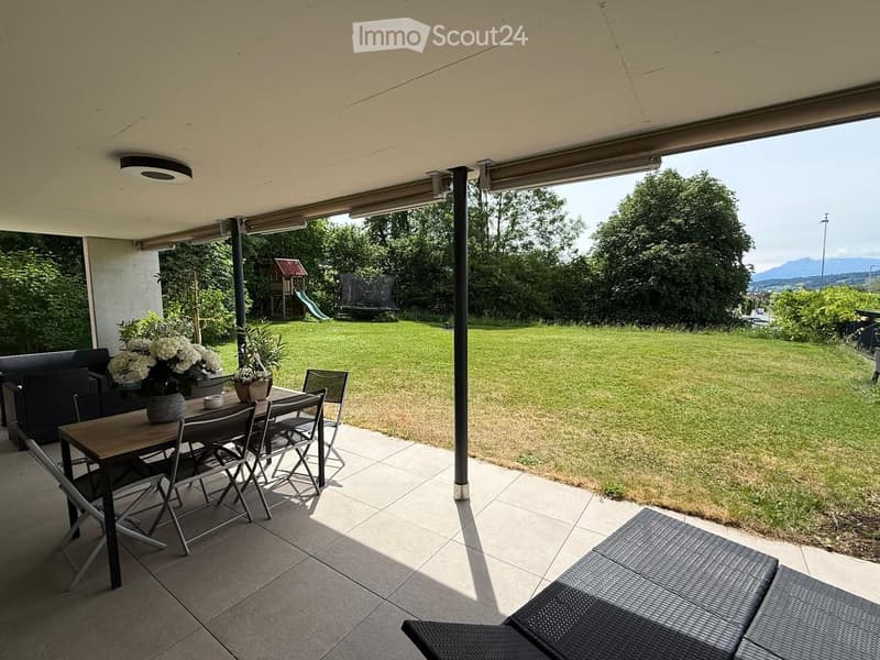 A spacious, covered balcony with a wooden table, chairs, a sofa, and flower pots, overlooking a well-maintained lawn and garden area with trees and a view of distant mountains.