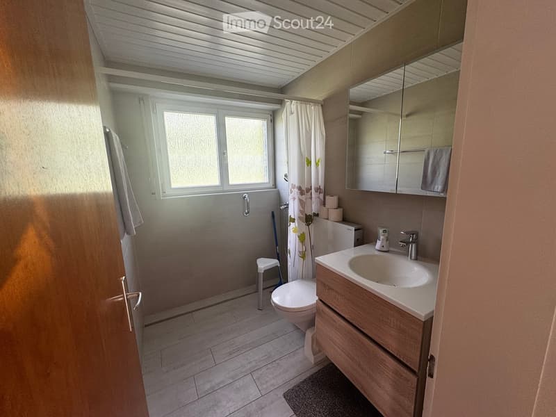 Bathroom. Modern bathroom with a wooden vanity, sink, mirror, toilet, and a shower area with a curtain. Includes a small stool and a towel on the cabinet.