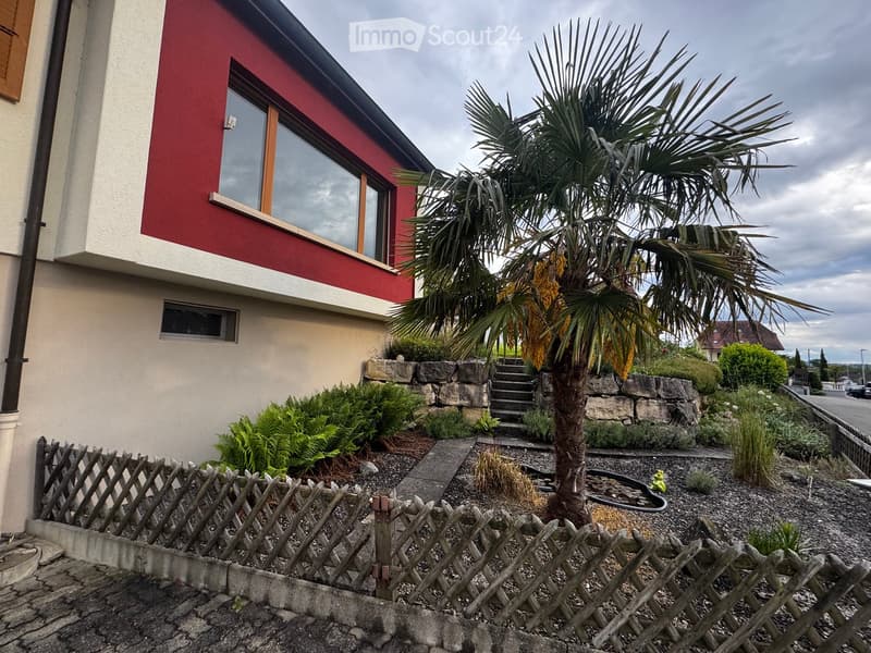 Garden / Outdoor Area. A two-story house with red and white exterior walls, large windows, and a well-maintained garden featuring a palm tree, shrubs, and a stone pathway leading to the entrance.