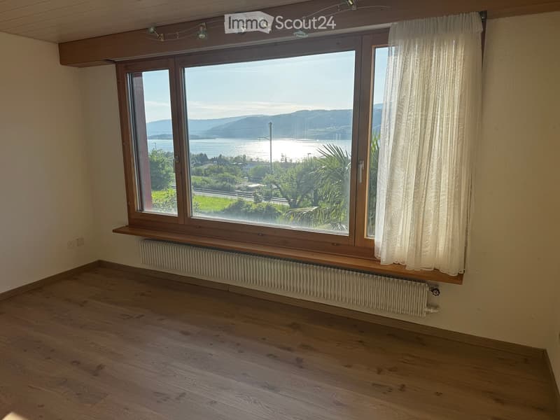 Unfurnished Room. Empty room with wooden flooring, large window offering a scenic view of a body of water and distant hills, radiator below the window, and a curtain on one side.