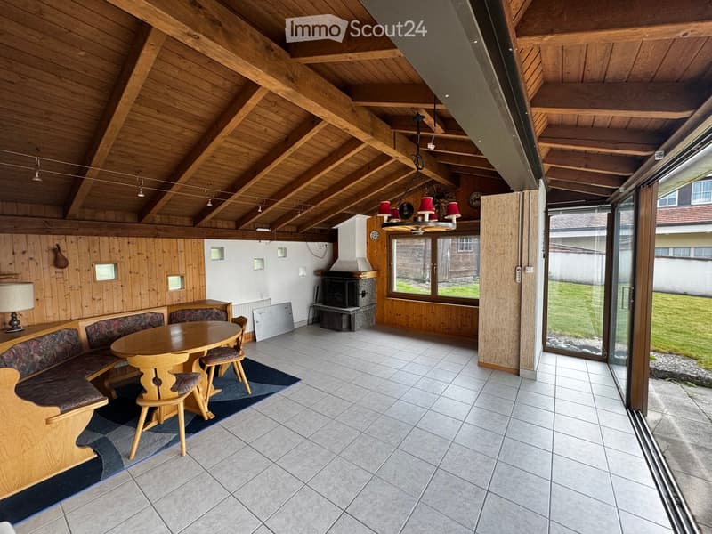 Furnished Room. Cozy living room with wooden beams, a round wooden table with chairs, a wooden bench, a fireplace, and large windows overlooking a green area.