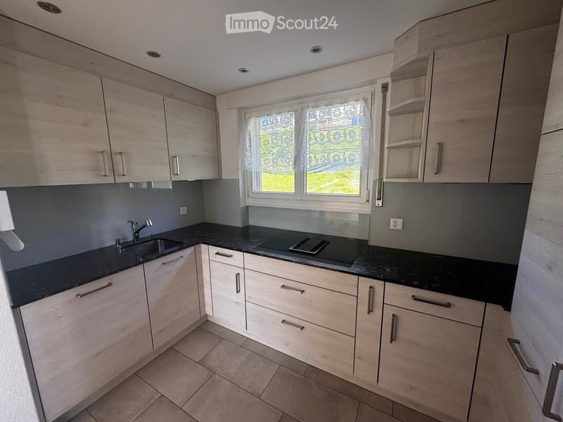 Kitchen. Modern kitchen with light wood cabinets, black countertops, a sink, and a stove. The space features a window with a decorative curtain and recessed ceiling lighting.
