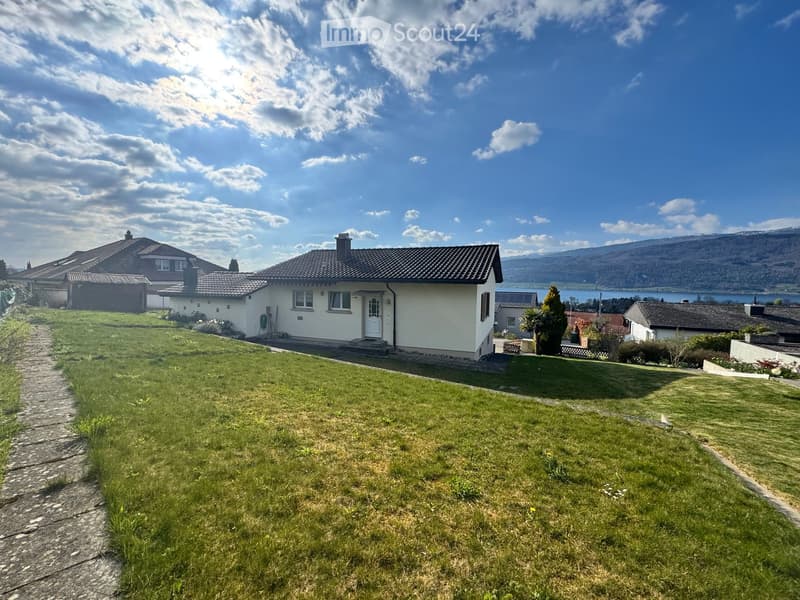 House. A single-story house with a tiled roof, white exterior, and a small front yard with grass and a stone pathway leading to the front door. The house is surrounded by other houses and a scenic view of a lake and mountains in the background.