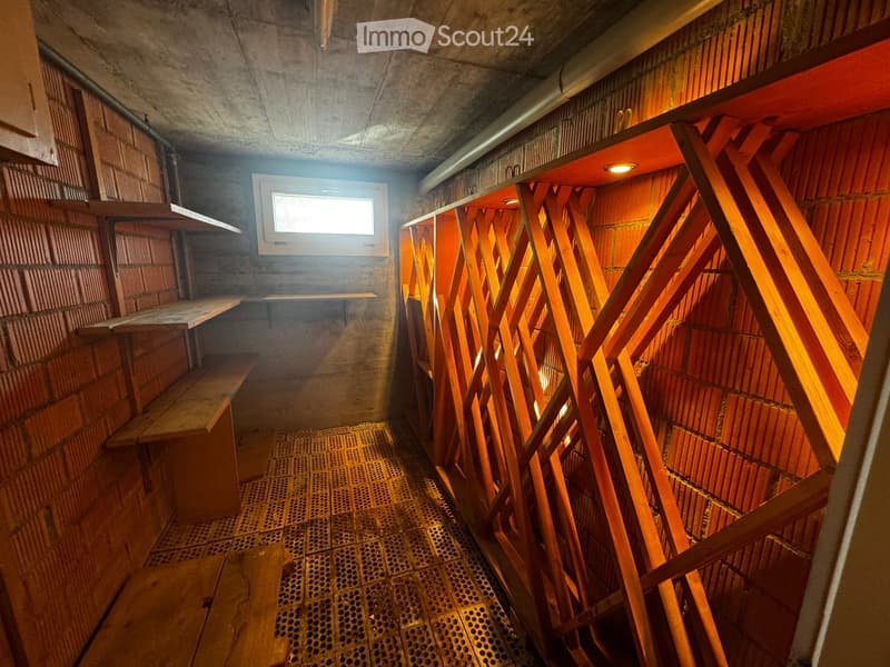 Corridor / Hallway. A narrow, dimly lit corridor with concrete walls, wooden benches, and a metal grated floor. A small window allows natural light to enter.
