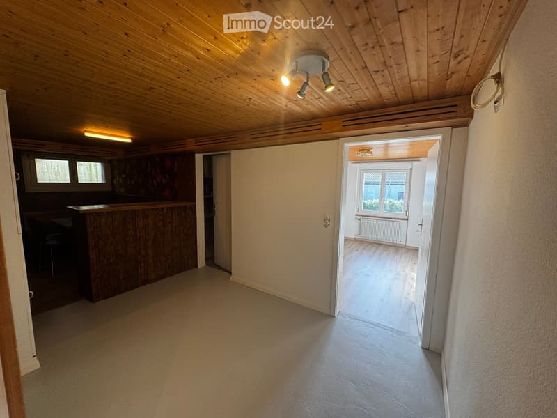 Corridor / Hallway. Empty hallway with wooden ceiling, light-colored walls, and a bar area visible to the left. The hallway leads to another room with a window and radiator.