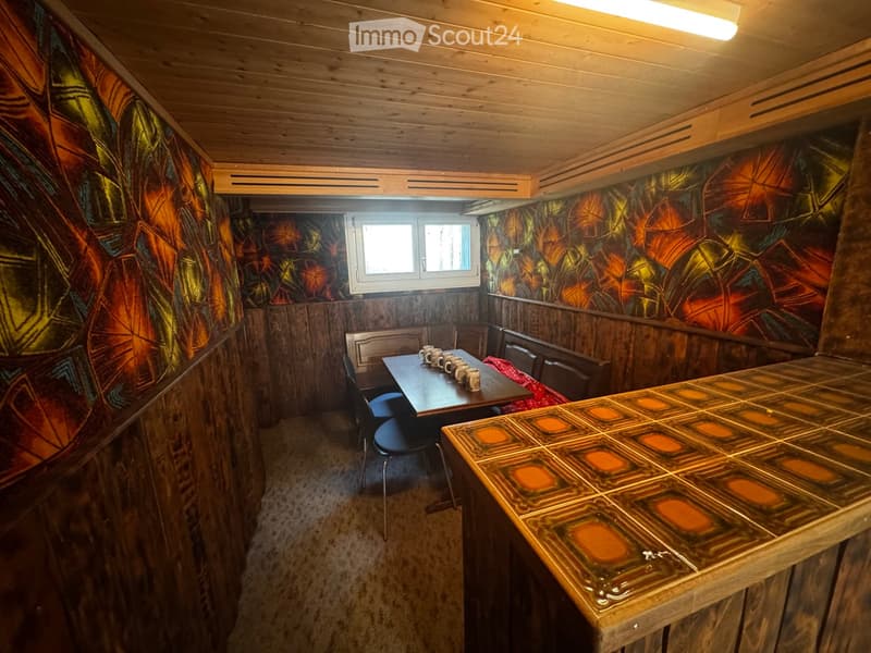 Furnished Room. A cozy wooden-paneled room with a table and chairs, colorful patterned wallpaper, and a window letting in natural light.