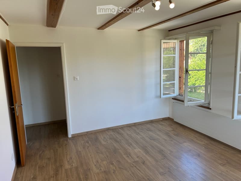 Empty room with wooden flooring, white walls, and large windows with open shutters. There is a visible radiator and an open doorway leading to another space.