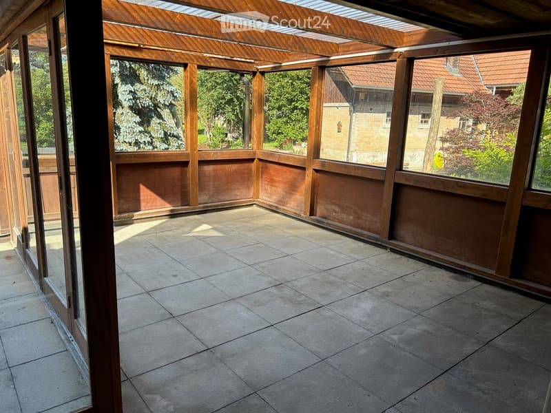 Empty wooden-framed balcony with tiled floor, large windows, and open sides, overlooking a garden and neighboring houses.