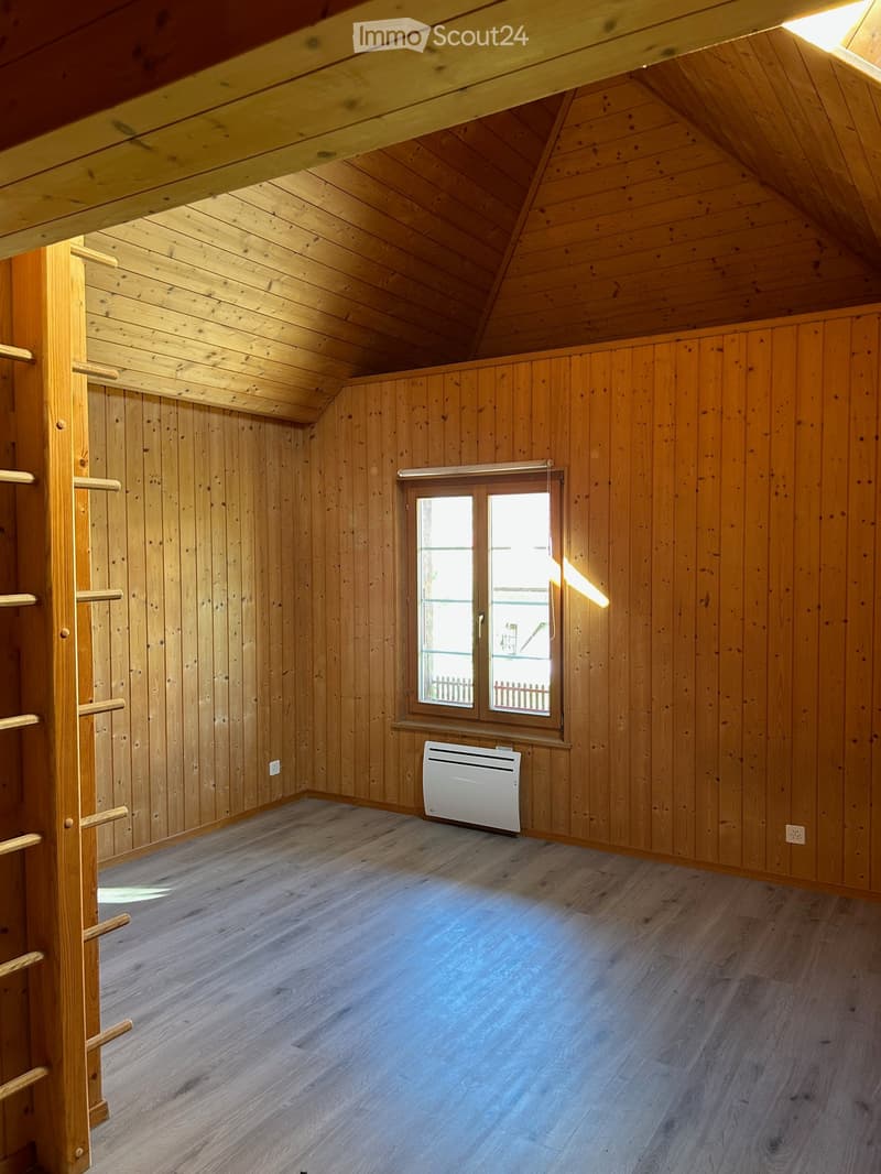 Empty attic room with wooden walls, ceiling beams, and a window. Features a white radiator and a wooden ladder leading to the loft area.