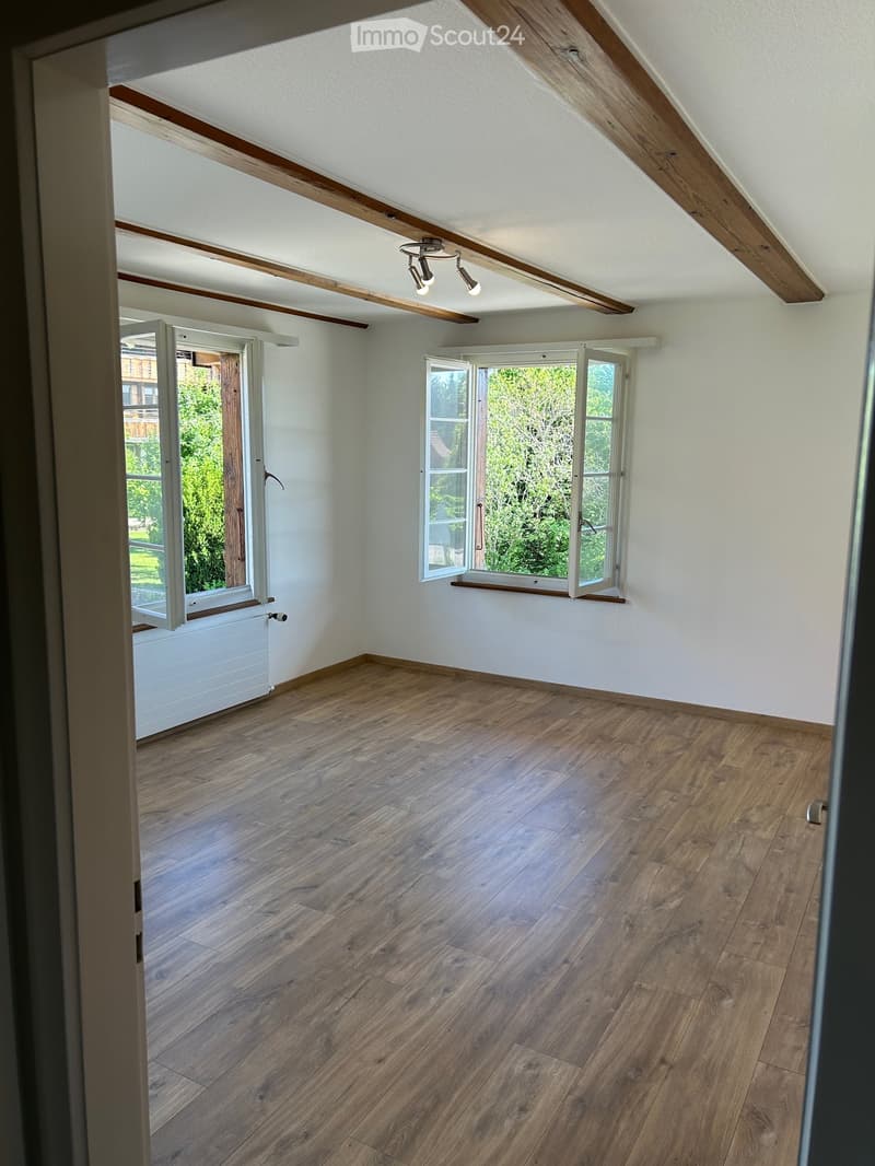 Empty attic room with wooden floors, white walls, exposed wooden beams, and large windows offering a view of greenery.