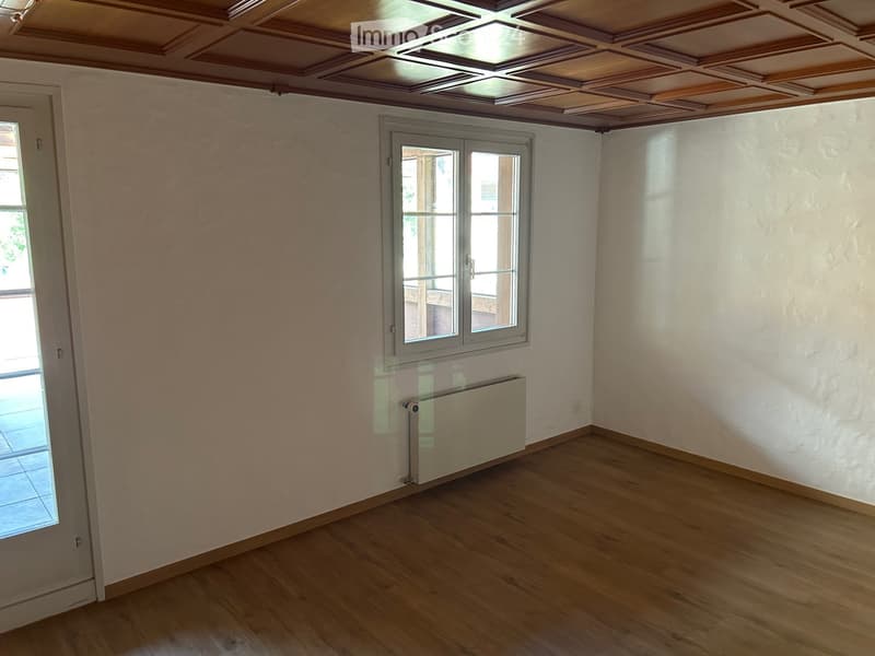Empty room with wooden flooring, white walls, and a radiator. Features a large window and a door leading to another space.