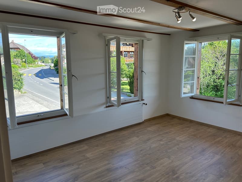 Empty room with wooden flooring, large windows, and exposed wooden beams on the ceiling. The room has a radiator under one window and offers a view of the street and surrounding greenery.