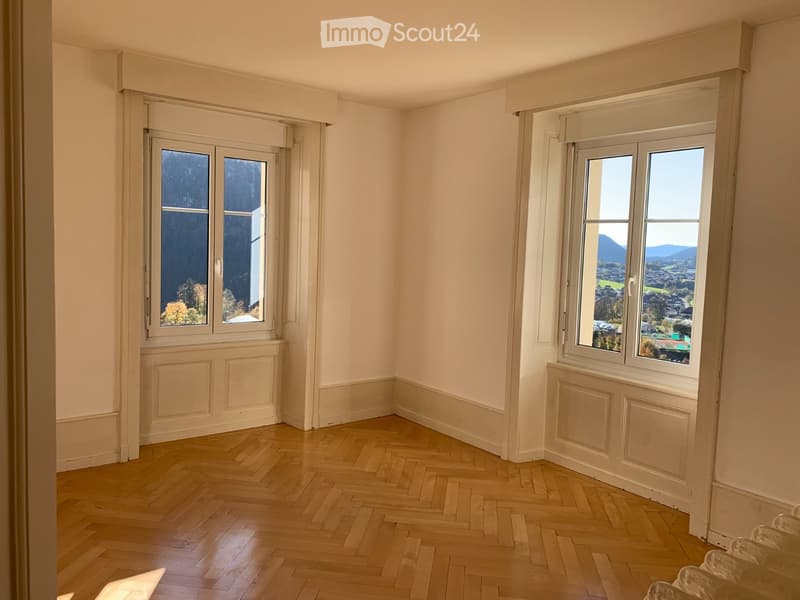 Empty room with wooden herringbone flooring, white walls, and large windows offering scenic views.