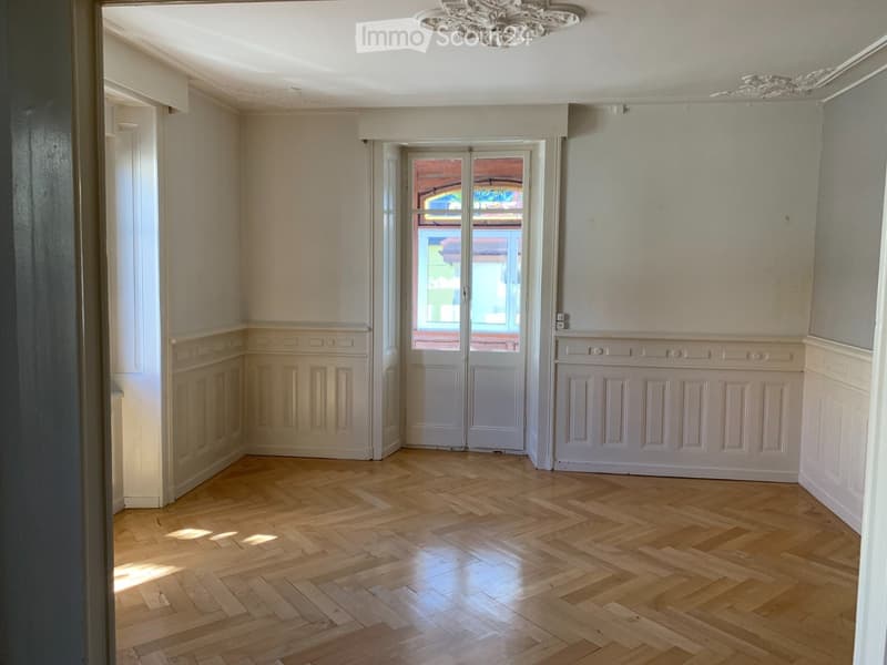 Empty room with wooden herringbone flooring, white walls, and a large window with double doors leading outside.