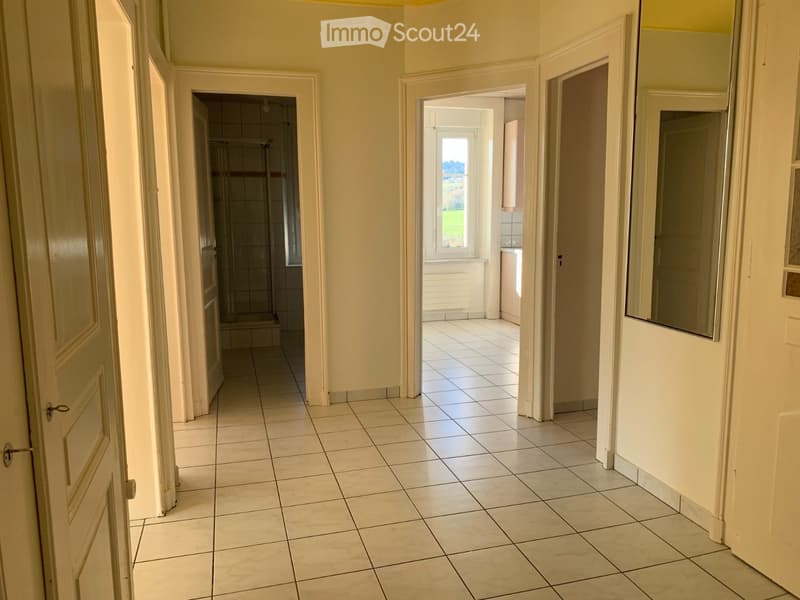 A clean, tiled hallway with multiple doors leading to different rooms, including a bathroom and a kitchen area visible through an open doorway.