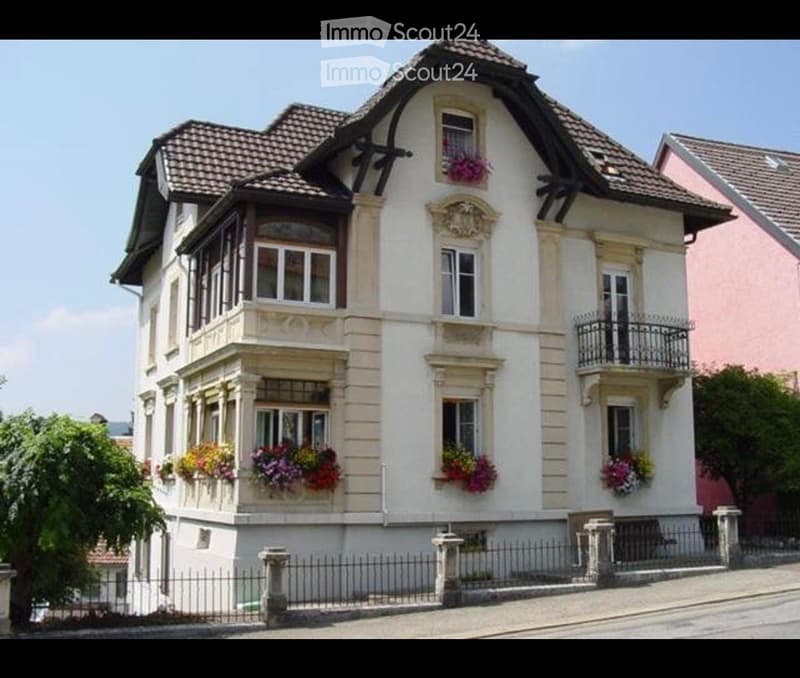 Two-story house with white facade, decorative balconies, and flower boxes. The roof is tiled, and the house features ornate architectural details.
