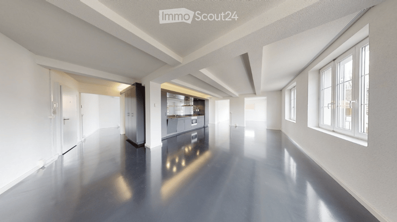 Open-plan unfurnished space with polished concrete floors, white walls, and large windows. The area includes a small kitchen area with cabinets and appliances, but no furniture is present.