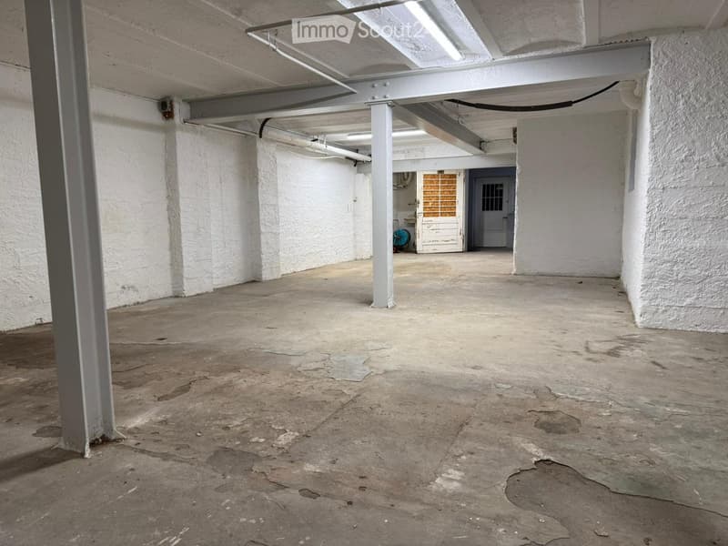 Empty, industrial-style room with concrete floor, white walls, exposed pipes, and a single door leading to another area.