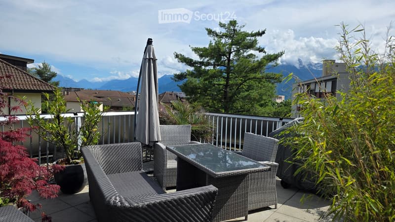 Balcony with wicker furniture, glass table, umbrella, and potted plants, overlooking a scenic mountain view.