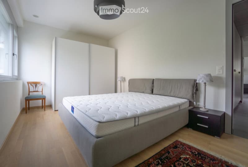 Modern bedroom with a large bed, nightstand, chair, wardrobe, and a patterned rug on the wooden floor.
