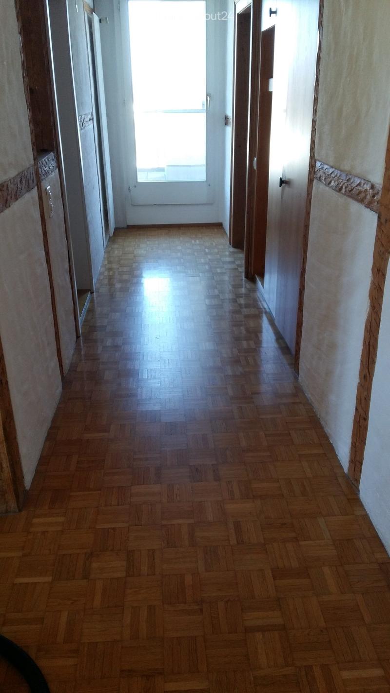 Empty hallway with wooden herringbone flooring, white walls, and doors on both sides, leading to a bright window at the end.