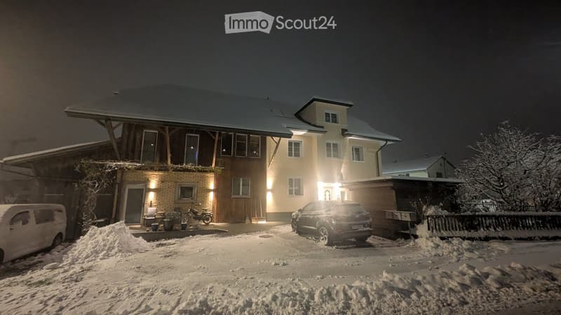 Two-story house with illuminated windows and a covered entrance, surrounded by snow-covered ground and parked vehicles.