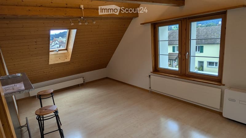Bright, unfurnished room with wooden walls, large windows, and a slanted wooden ceiling. Two stools and a small table are present.