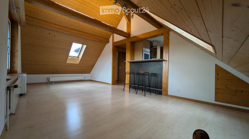 Bright, open loft-style room with wooden beams, exposed wooden ceiling, and skylight windows. Features a small kitchen area with a countertop and three stools, a radiator, and a wooden floor.