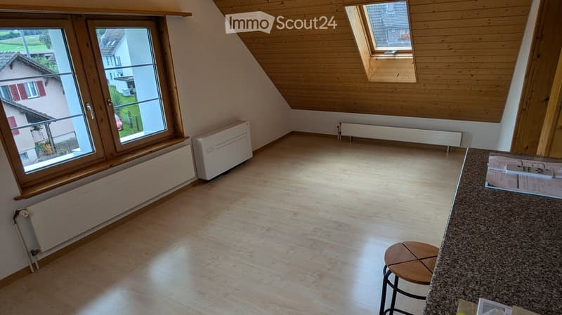 Empty attic room with wooden slanted ceiling, large windows, light-colored wooden floor, radiator, and a small wooden stool near a counter.