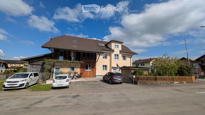 Two-story house with a combination of brick and plaster exterior, multiple windows, and a wooden garage section. There are three parked cars in front of the house, and the surrounding area includes a small garden and neighboring houses.