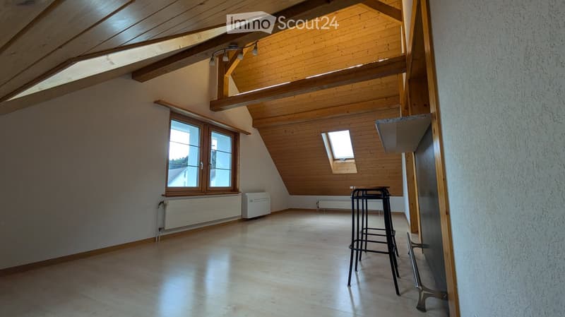 Empty attic room with wooden slanted ceilings, large windows, and a small table and chairs.