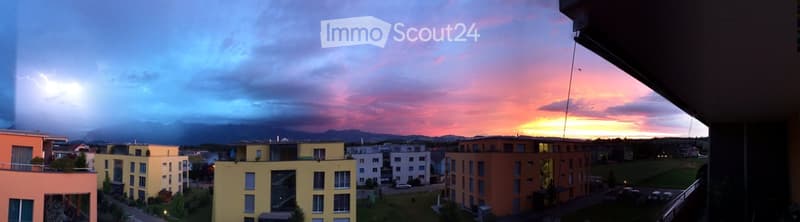 Balcony view of a residential area at sunset, with modern apartment buildings, green spaces, and distant mountains.