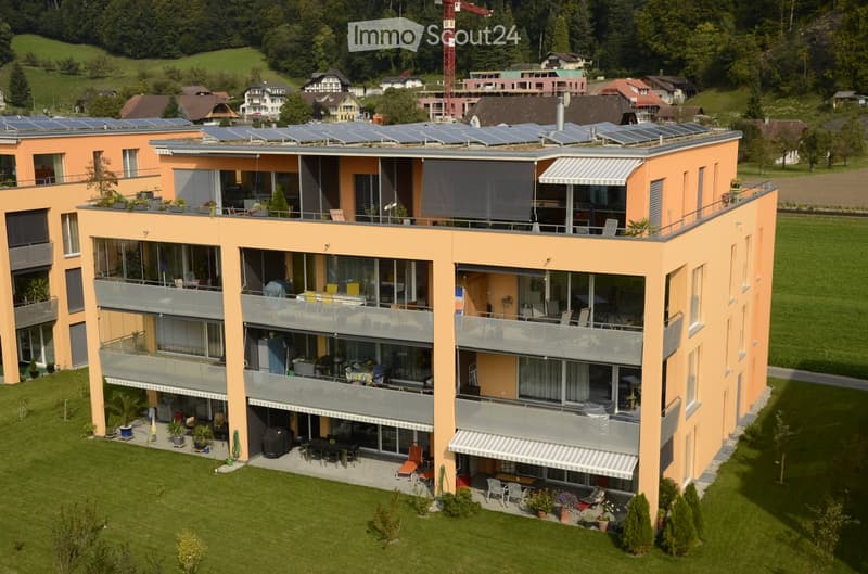 A modern, multi-story building with orange exterior walls, large windows, balconies, and solar panels on the roof. The building is surrounded by a well-maintained lawn and garden area with outdoor furniture and potted plants.