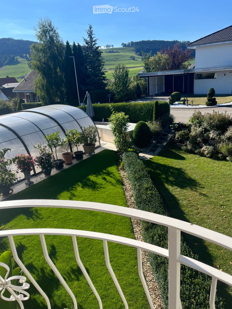 Well-maintained garden with green lawn, trimmed hedges, potted plants, and a greenhouse. The view includes a scenic countryside with hills and trees.