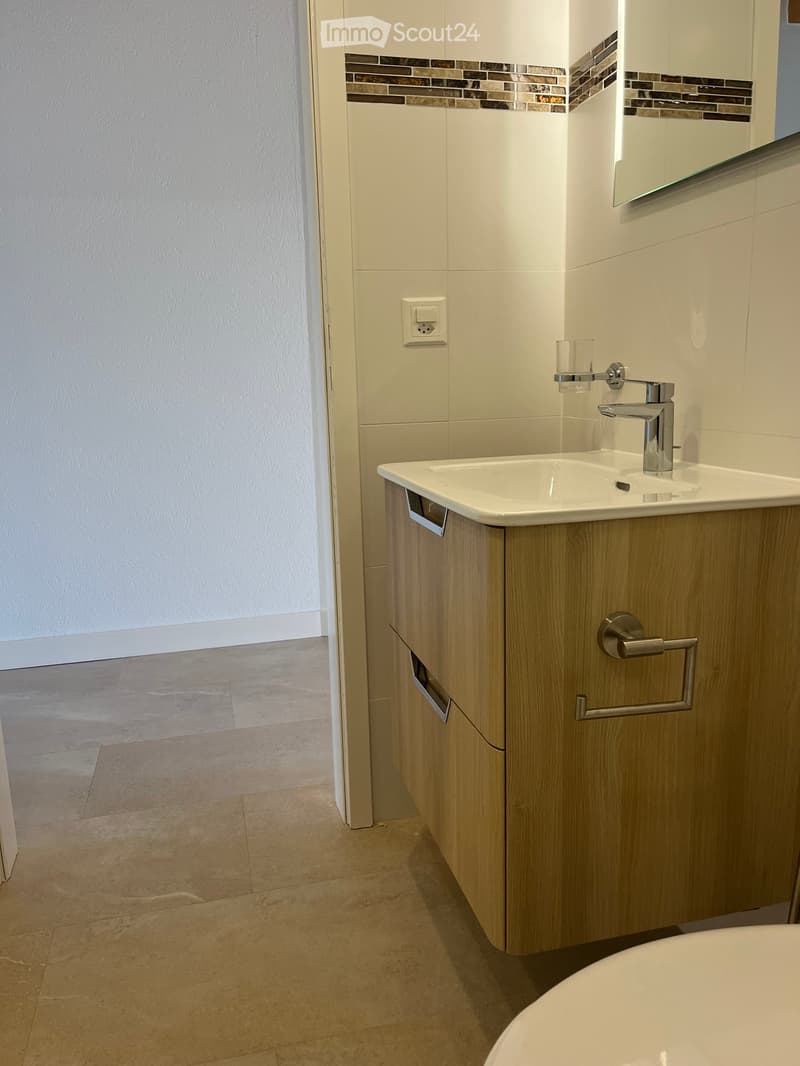 Modern bathroom with a white sink, chrome faucet, wooden cabinet, and a mirror above the sink. The walls are tiled with a decorative border, and the floor is tiled.