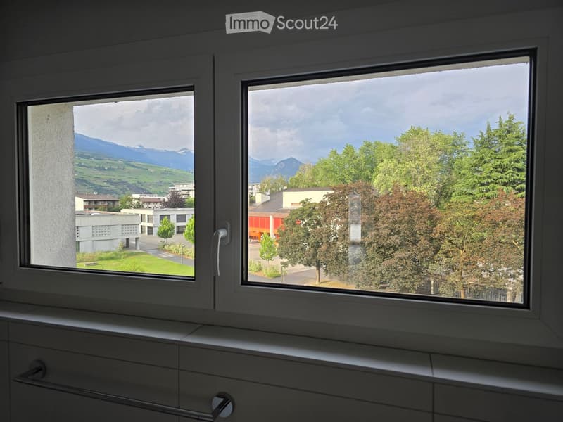 Bathroom with large windows offering a scenic view of mountains, trees, and buildings. The windows are framed in white and have a modern design.