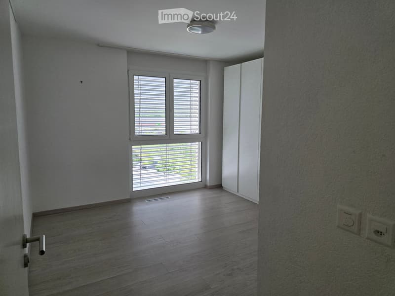 Empty room with wooden flooring, white walls, large window with blinds, and a closed white door.