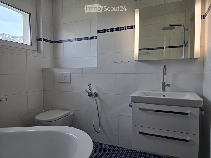 Modern bathroom with a bathtub, toilet, sink, and showerhead. Features a large mirror, sleek fixtures, and a window with a scenic view.