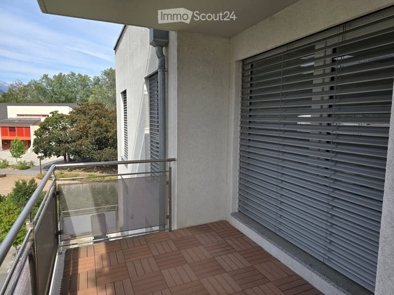 Balcony with wooden decking, metal railing, and closed blinds on the windows.