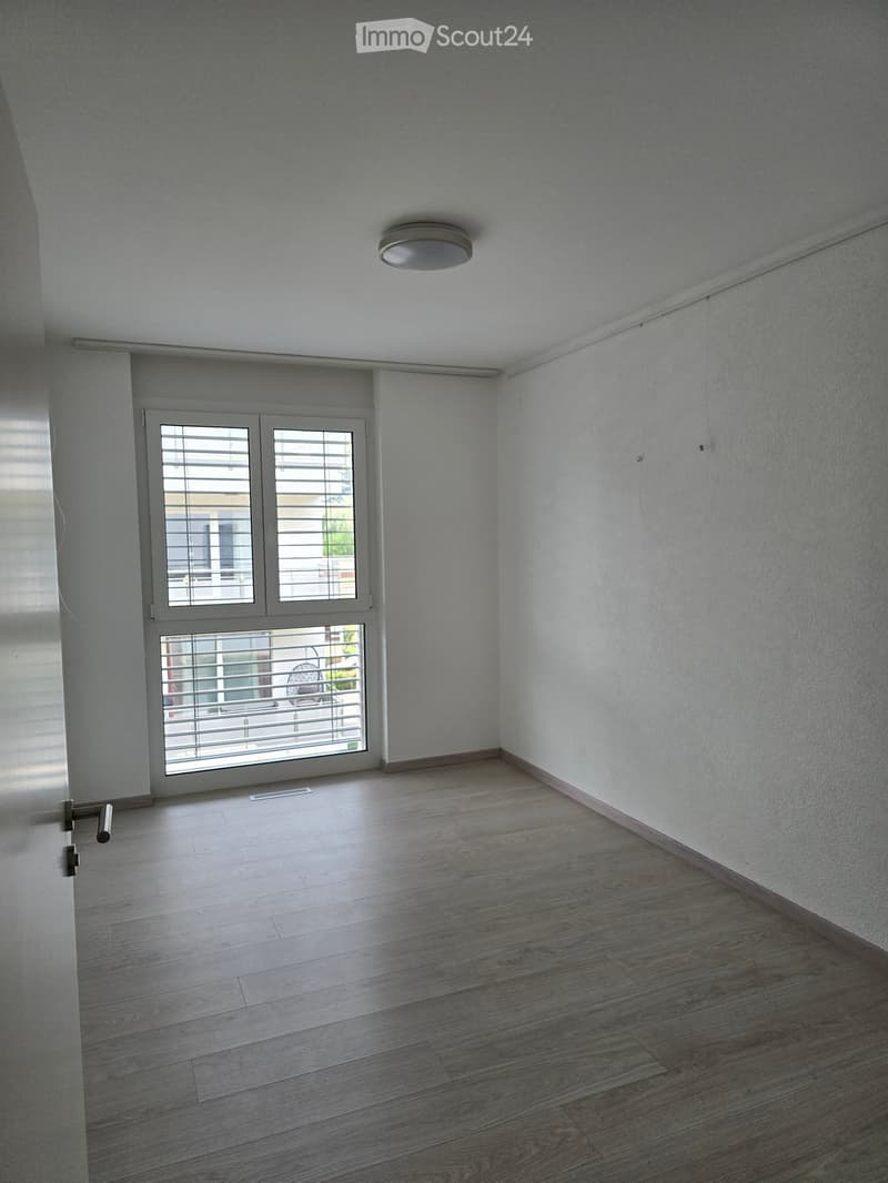 Empty room with light grey walls, wooden flooring, and a large window with blinds. The room has a modern ceiling light fixture and a door with a handle visible on the left side.