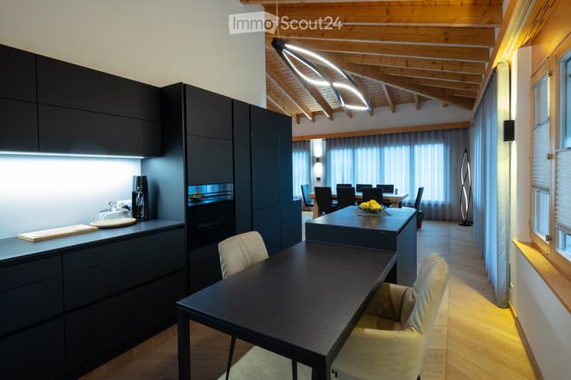 Modern kitchen and dining area with dark cabinets, a central island, and a dining table with chairs. The space features wooden beams on the ceiling and large windows with curtains.