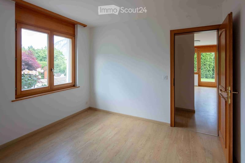 Empty room with light wooden flooring, white walls, and large windows with wooden frames. The room has an open doorway leading to another space with similar flooring and windows.
