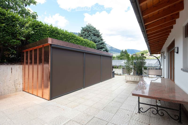A modern outdoor patio area with a wooden table, a copper privacy screen, and a view of surrounding greenery and mountains.