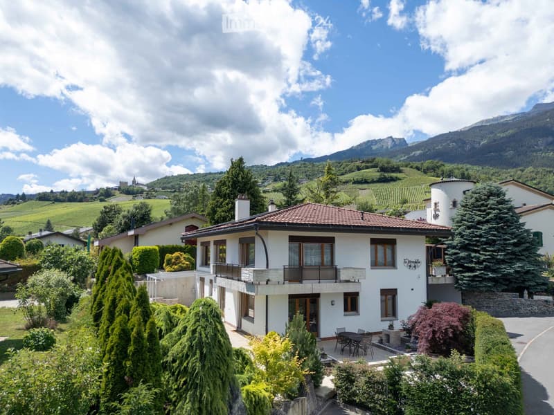A two-story white house with a red-tiled roof, large windows, and a balcony overlooking a scenic valley with vineyards and mountains in the background.